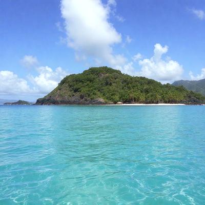 The primary subject of the image is a small island surrounded by a large body of water. The island is located in the middle of the ocean, and it appears to be a tropical paradise. The setting/background features a clear blue ocean, with a few clouds scattered in the sky. The visual style of the image is a photo, capturing the beauty of the island and its surroundings. The colors and mood of the image are vibrant and serene, with the blue ocean and the lush green island creating a sense